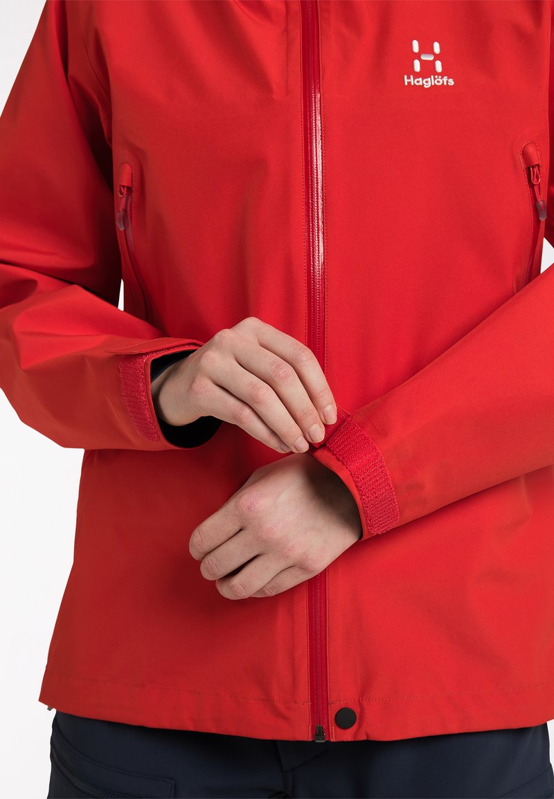 Červená voděodolná bunda s předním zipem a suchými zipy na manžetách, s logem na hrudi a dvěma bočními zipovými kapsami.