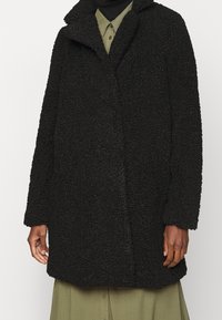 Cappotto nero e texturizzato realizzato in materiale morbido, caratterizzato da un colletto a punta e una vestibilità rilassata. Sono visibili le maniche lunghe e il dettaglio di chiusura frontale.
