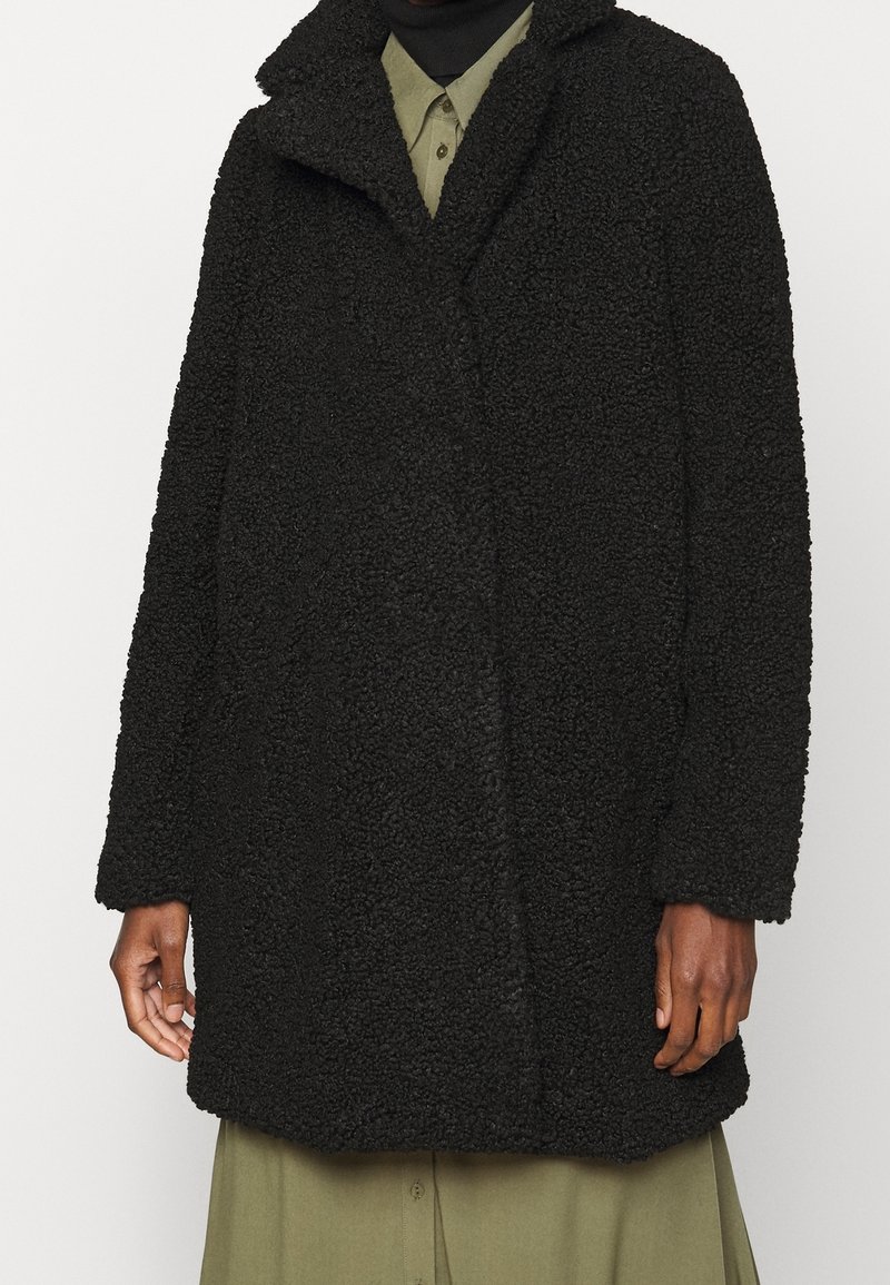 Cappotto nero e texturizzato realizzato in materiale morbido, caratterizzato da un colletto a punta e una vestibilità rilassata. Sono visibili le maniche lunghe e il dettaglio di chiusura frontale.