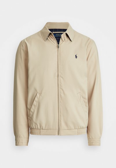 Veste zippée beige clair avec un col, des poches latérales, une taille élastique et un petit logo bleu marine sur la poitrine. Texture lisse.
