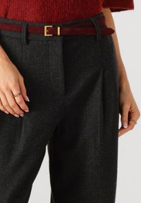 Donkergrijze visgraat broek met een relaxte pasvorm, voorzien van een bordeauxrode suède riem en een gouden metalen gesp. Hand op de tailleband geplaatst.