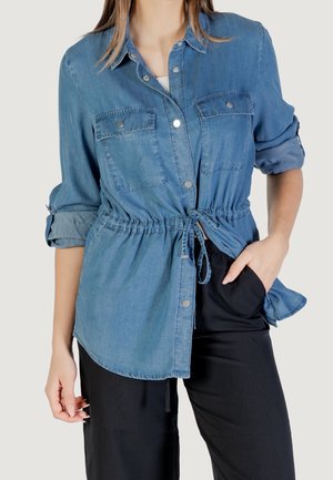 Vrouw die een lichtblauw spijkershirt draagt met opgerolde mouwen en een koord aan de voorkant, gecombineerd met een zwarte broek, hand in de zak.