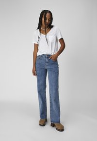 Hvit kortermet t-skjorte med lomme, kombinert med høydejusterte blå jeans. Modellen har på seg brune platåklokker og står mot en grå bakgrunn.