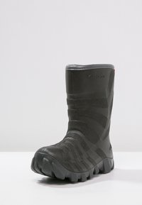 Schwarzer Gummistiefel mit strukturierten Oberflächen, erhöhten Streifen und einer dicken, genoppten Sohle. Abgerundete Zehenpartie und leicht erhobene Ferse.