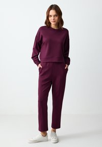Pruimen kleurige trainingspak gemaakt van zacht materiaal, met een cropped top met ingezakte schouders en taps toelopende broek met zijzakken. Witte sneakers.
