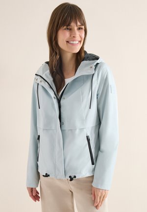 Usměvavá žena v světle modré kapuci s černými šňůrkami a kapsami na zip, stojící před jednobarevným béžovým pozadím.