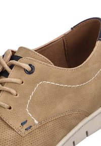 Zapato casual de ante marrón con cordones beige, detalle de costura blanca y suela de goma blanca, mostrando vista lateral y superior en primer plano.