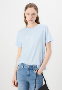 Heledad hallblue T-särk, lühikeste varrukatega ja ümmarguse kaelusega, millel on valge trükitud logo. Koos hallide siniste teksade ja väikese musta käekotiga.