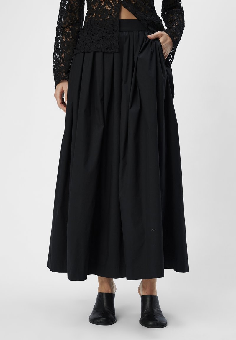 Svart, veckad midikjol med hög midja och sidofickor, tillverkad av ett mjukt tyg. Bärs med svarta slip-on-skor.