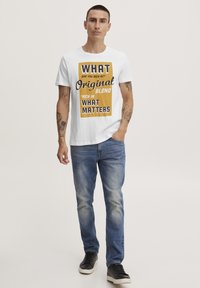 Valge puuvillane t-särk, millel on paksud kollased ja mustad kirjatsed, kombineeritud siniste teksapükste ja mustade tennistega. Lihtne disain, vabaaja lõige.