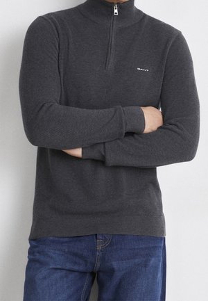 Uomo con le braccia incrociate che indossa un maglione grigio scuro con zip a quattro quarti con logo GANT e jeans blu su uno sfondo chiaro e uniforme.