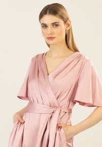 Rosenfarbendes Satin-Wickelkleid mit kurzen Ärmeln, V-Ausschnitt, gerafften Details und gebundenem Tailleneinsatz. Zwei seitliche Taschen sind sichtbar. Glatte Textur.