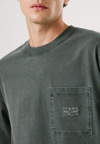 Camiseta de algodón gris oscuro con cuello redondo, mangas cortas y un bolsillo en el pecho que presenta una etiqueta de logo bordada. Tejido suave y texturizado.