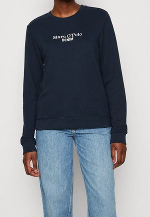 Persoon draagt een marineblauwe longsleeve trui met de tekst "Marc O’Polo DENIM" en lichtblauwe spijkerbroek, staand tegen een effen achtergrond.