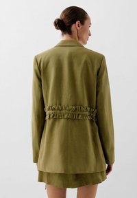 Olive green blazer with a ruffled accent at the waist, featuring long sleeves and a sleek design, shown from the back.