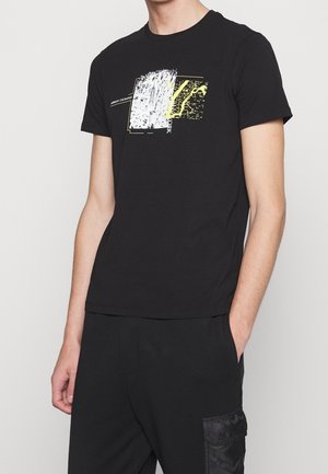 Homme portant un t-shirt noir à manches courtes avec un graphisme abstrait blanc et jaune et un pantalon noir avec une poche latérale, debout devant un fond blanc.