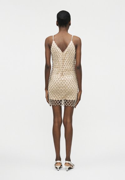 Γυναίκα όρθια με την πλάτη στην κάμερα, φορώντας μπεζ φόρεμα slip καλυμμένο με ασημένιο πλεκτό στρώμα και λευκά σανδάλια με τακούνι kitten.