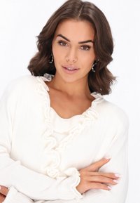 Fehér bordás pulóver ráncos nyakkivágással és mandzsettákkal, amely puha tapintású és hosszú ujjú. A modell karba tett kézzel pózol.