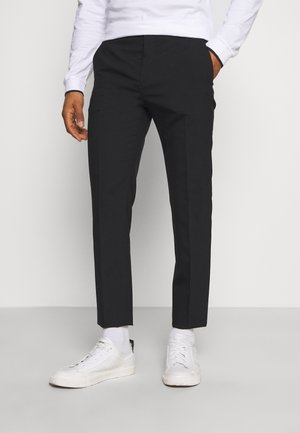 Mann trägt schlanke schwarze Hose, weißes Langarmhemd, weiße Socken und weiße Turnschuhe, steht mit einer Hand in der Tasche.