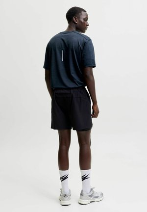 Homme portant un t-shirt bleu foncé, un short noir, des chaussettes blanches avec un motif noir et des baskets grises, debout et regardant sur le côté.
