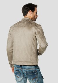 Béžová semišová bunda so štruktúrovaným dizajnom, vybavená prešívanými ramennými detailmi a priliehavým strihom. Kombinovaná s modrými džínsami.