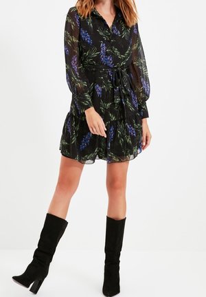 Schwarzes, transparentes Kleid mit grün-blauem Blumenmuster, langen Ärmeln und tailliertem Gürtel. Kombiniert mit kniehohen schwarzen Wildlederstiefeln.