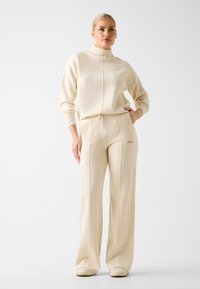 Beige matching set featuring a zip-up high-neck top and wide-leg pants, crafted from soft fabric with logo detail and visible stitching.