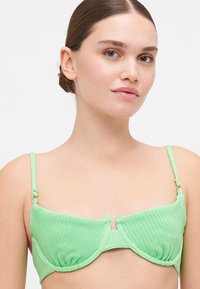 Grønn teksturert bikini-topp med zigzag-mønster, med justerbare stropper og detaljer med frontklips. Glatt, strukturert design.