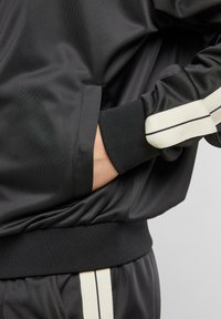 Veste de sport noire en tissu lisse, avec des poignets côtelés et des poches latérales. Les manches présentent des rayures contrastantes couleur blanc cassé.