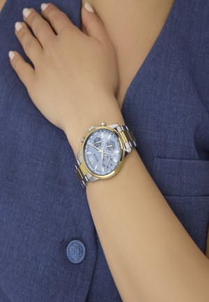 Montre en métal argenté et doré avec un cadran bleu, trois sous-cadrans et des marqueurs en chiffres romains, portée sur une main reposant sur un tissu bleu.