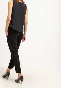Top nero senza maniche con apertura a chiave sul retro, abbinato a pantaloni neri a sigaretta e tacchi alti neri, caratterizzato da una superficie liscia e una vestibilità sartoriale.