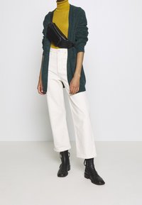 Vihreä villatakki, sinapinkeltainen poolopaita, valkoiset leveälahkeiset housut ja mustat nilkkurit. Vyölaukku on teksturoitu ja musta. Siisti, minimalistinen design.