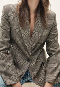 Bruine geruite blazer met een notched revers, sluiting met één knoop en iets oversized mouwen. Gecombineerd met een licht denim outfit.