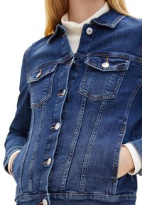 Modrá džínová bunda s předním zapínáním na knoflíky, dvěma kapsami na prsou a strukturovaným střihem s prošitími a stříbrnými knoflíky.