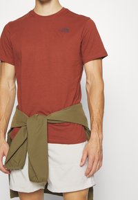 Camiseta de algodón roja con manga corta y un pequeño logo, combinada con pantalones cortos claros. Una prenda de color verde oliva está atada alrededor de la cintura.