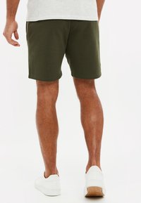 Homme portant un short vert olive, des baskets blanches et une chemise gris clair, debout de dos sur un fond blanc uni.