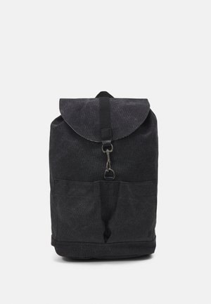 Sac à dos en tissu noir avec rabat avant sécurisé par une fermeture métallique et deux poches avant, présenté sur un fond blanc uni.