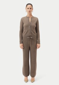 Brunt stickat set med ett strukturerat mönster, bestående av en knappad kofta och byxor med dragsko i midjan. Spetsiga skor fullbordar outfiten.