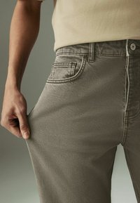 Grijze denim broek met een aansluitend ontwerp, zichtbare stiksels en een voorzak. Een hand trekt aan de stof aan de zijkant voor demonstratie.