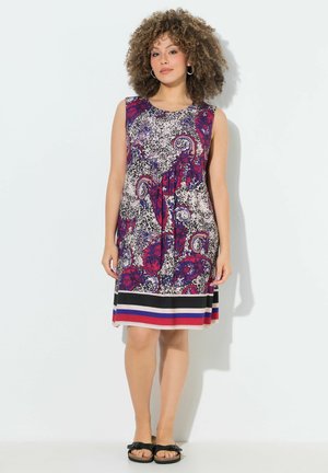 Kvinde med krøllet hår bærer en ærmeløs knælængde kjole med rød, lilla paisley og leopardprint, stribet kant og sorte sandaler.