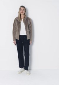 Kurtka z futerka ekologicznego w jasnobrązowym kolorze z kołnierzem i przednimi kieszeniami, zestawiona z ciemnymi dżinsami i białymi trampkami, na jednolitym tle.