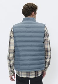 Gilet rembourré gris avec un design à cannelures verticales, col montant et sans manches, porté sur une chemise en flanelle à carreaux. Présente une texture lisse.