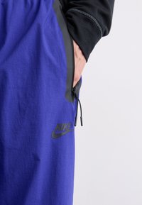 Pantalones cortos deportivos morados con un bolsillo lateral negro con cremallera, acentuados con el logo de Nike en negro. Fabricados con tela ligera.