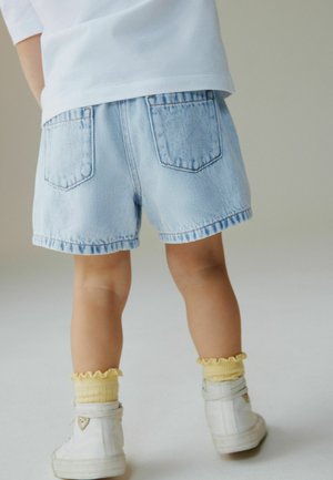 Copil purtând pantaloni scurți din denim albastru deschis, șosete galbene cu volane, adidași albi și o cămașă albă, stând în fața unui fundal simplu, deschis la culoare.