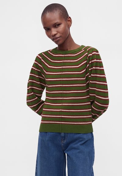 Personne aux cheveux courts portant un pull rayé vert, rouge et blanc et un jean bleu, debout devant un fond uni.