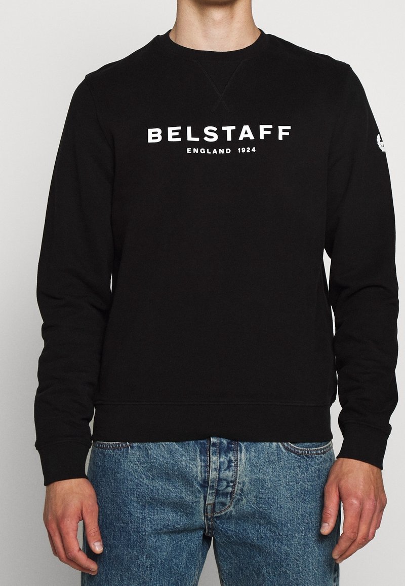 Personne portant un sweat-shirt noir avec un texte blanc "BELSTAFF England 1924" et un jean bleu, debout devant un fond clair uni.