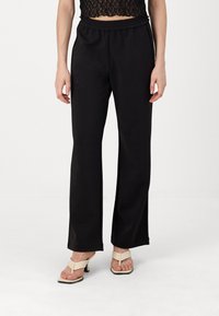 Μαύροι, φαρδιοί παντελόνες με ελαστική μέση, λεία υφή και πλαϊνές σχισμές, συνδυασμένοι με μπεζ τακούνια.