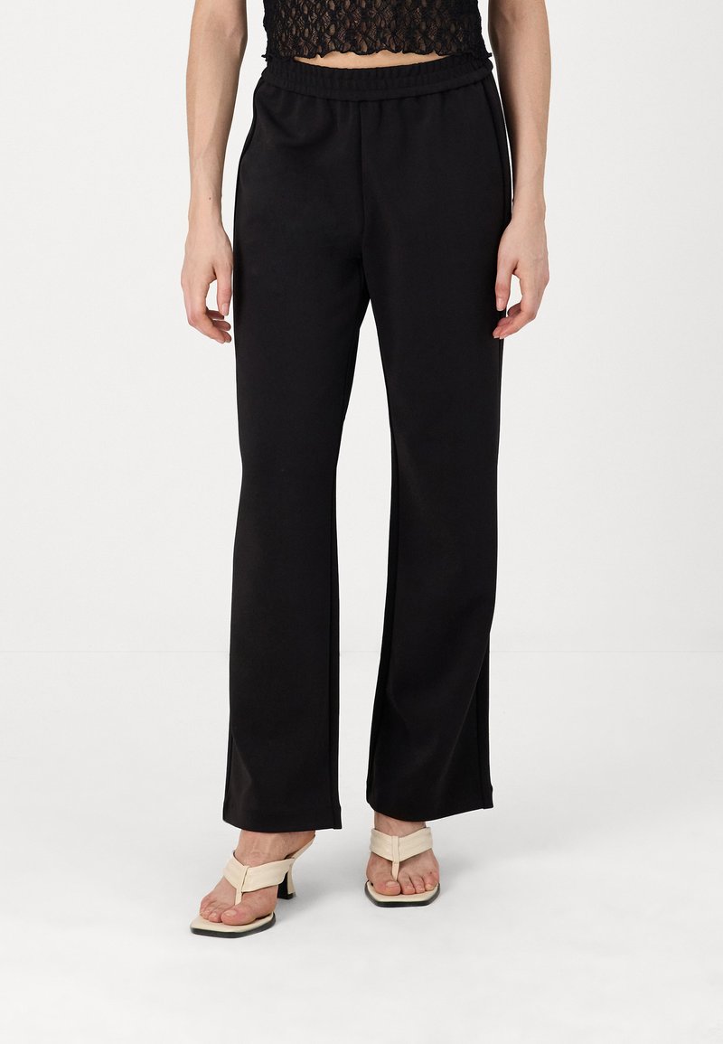 Μαύροι, φαρδιοί παντελόνες με ελαστική μέση, λεία υφή και πλαϊνές σχισμές, συνδυασμένοι με μπεζ τακούνια.