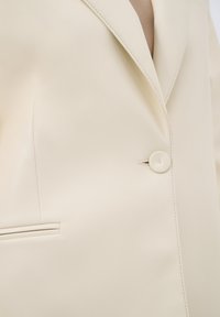 Blazer color crema realizzato in tessuto liscio e lucido, con chiusura a un bottone e rever a punta, dotato di dettaglio con tasca laterale.