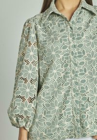 Blusa in pizzo verde chiaro con motivo floreale, chiusura a bottoni e maniche a sbuffo, che mette in risalto ricami dettagliati e una texture traspirante.
